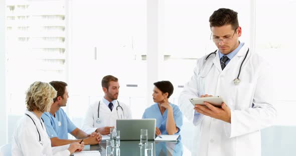 Thoughtful Doctor Using Tablet with Staff Talking Behind Him alt