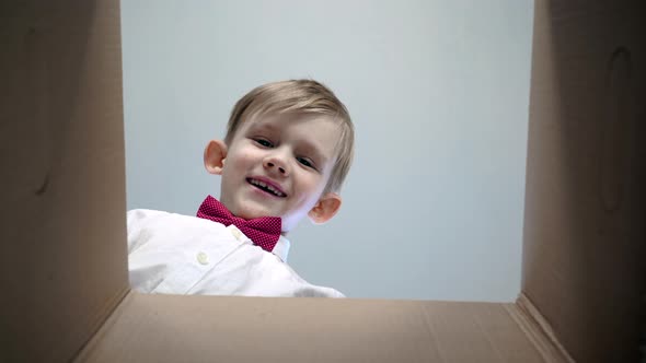 Happy Little Blond Boy in a White Shirt with a Red Bow Tie Opens a Cardboard Box and Smiles