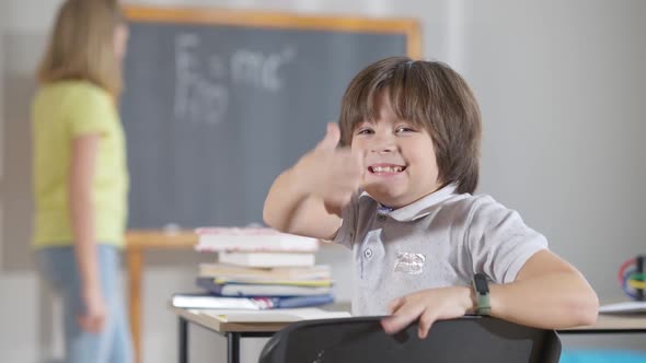Cute Excited Schoolboy Turning To Camera Showing Thumb Up with Blurred Classmate Writing on School alt