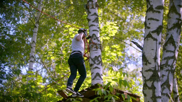 A Man in Helmet Carefully Walks on the Rope Suspended in the Air Between Trees in the Forest alt