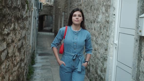 Pretty young woman with brunette hair in denim overalls walks and smiles in old town in Montenegro alt