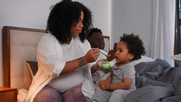 Young African American Mother Feeding Cute Son with Spoon As Blurred Father Reading Lying in Bed at alt
