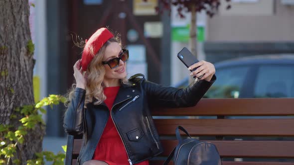 Young Woman in Stylish Clothes Taking a Selfie Sitting on a Park Bench alt