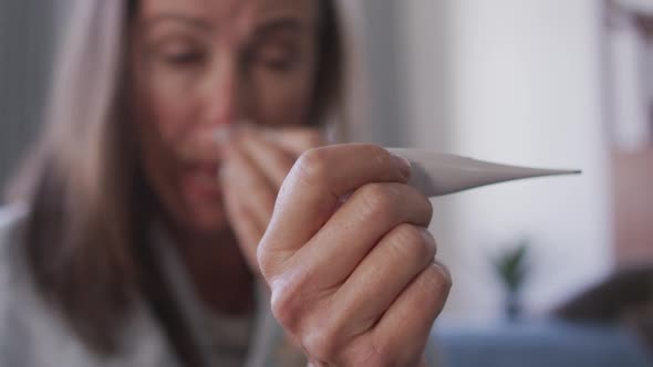 Woman holding a thermometer while blowing her nose at home alt