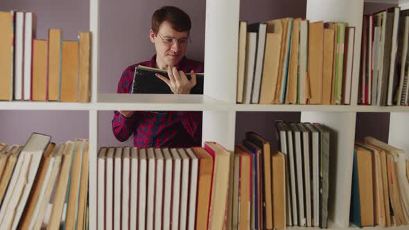A Man Wearing Glasses is Reading a Book in the Library Behind the Bookshelves alt