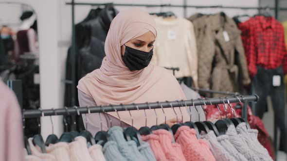 Young Arab Woman Shopper in Medical Protective Mask Chooses Clothes in Clothing Store Retail Sales alt