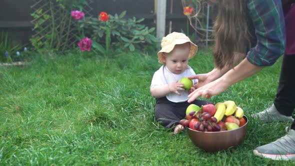 Small Newborn Child in Summer Panama Hat Sit on Grass Barefoot in Bib with Big Bowl of Fresh Fruit alt