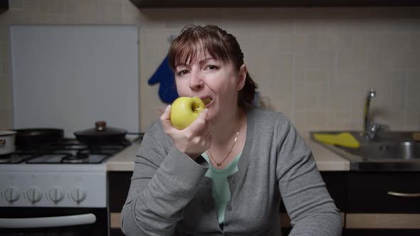 Woman Sits in the Kitchen Attentively with Excitement Looks at the Camera and Eats an Apple in the