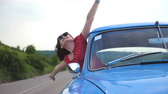 Young Smiling Girl in Sunglasses Leaning Out of Vintage Car Window and Enjoying Trip. Happy alt