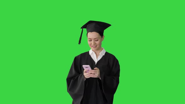 Smiling Female Graduate in Mortarboard Sharing Happy News on Her Phone While Walking on a Green alt