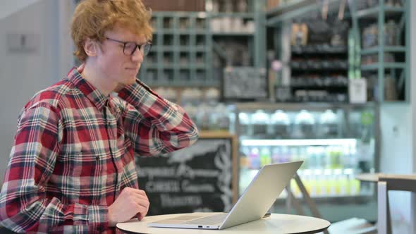 Young Redhead Man with Neck Pain Using Laptop in Cafe  alt