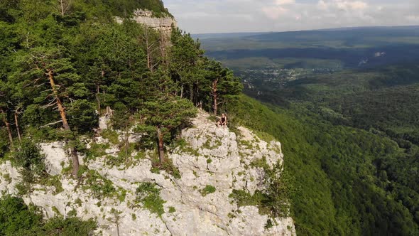 A Long-haired, Thin Man in a Black T-shirt and Shorts Sits on the Edge of a Cliff and Looks Out at alt