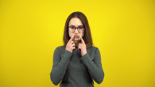 A Young Woman Makes a Cheerful Smile on Her Face with Her Fingers on a Yellow Background. Woman with alt