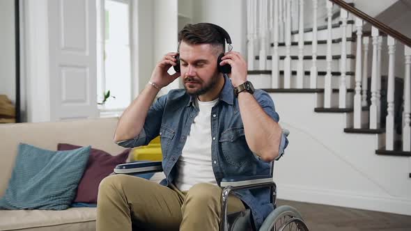 Bearded Guy in Wheelchair which Putting on Earphones and Listening Relaxing Music at Home alt