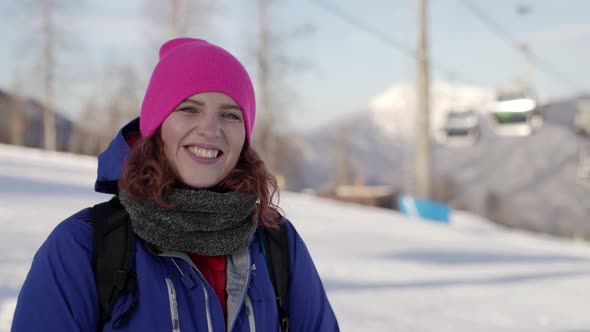 Portrait of a Woman in a Pink Hat and Blue Jacket Standing on the Background of Ski Lifts Smiling alt