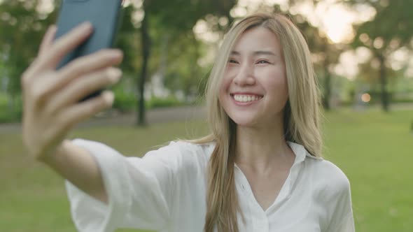 Asian woman taking selfie photo on smartphone at a public park during sunset.