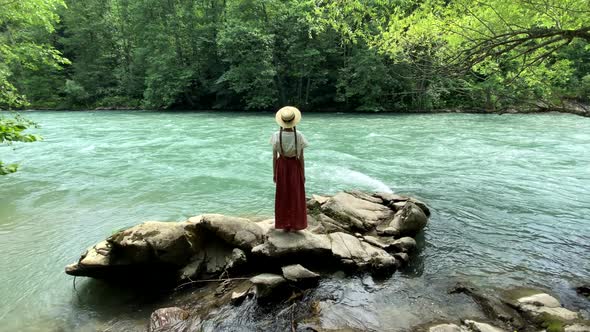Young vintage woman in dress and straw boater hat stand near rippling mountain river, back view