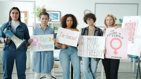 Group of Women with Feminist Posters Standing Indoors and Looking at Camera with Serious Faces alt