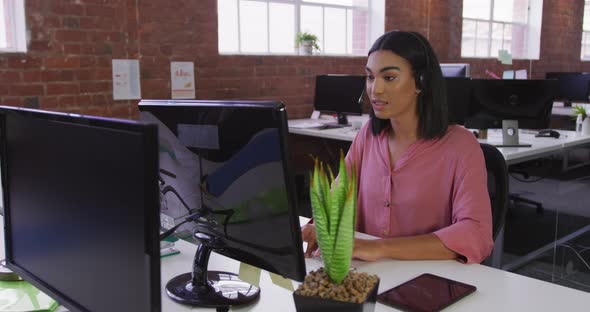 Mixed race businesswoman wearing headset sitting at desk having video call alt