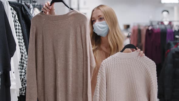 Young Girl in Medical Mask Choose Clothes in Store Woman Consumer Make Choice Between Two Sweaters alt