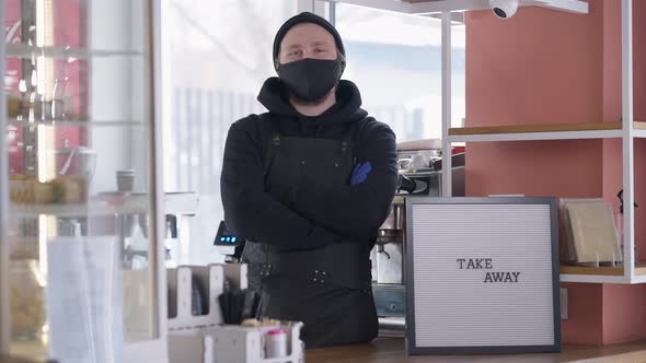 Confident Caucasian Young Man in Covid Face Mask Crossing Hands Looking at Camera Standing at Cafe alt