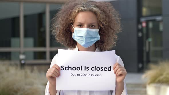 Schoolgirl Wearing a Medical Mask Holds a Sign SCHOOL IS CLOSED DUE TO COVID19 VIRUS alt