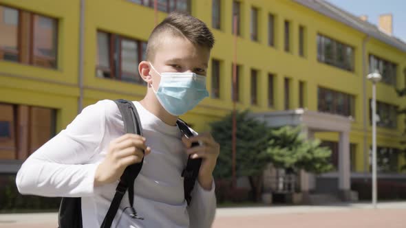 A Caucasian Teenage Boy Puts on a Face Mask and Looks at the Camera  a School in the Background