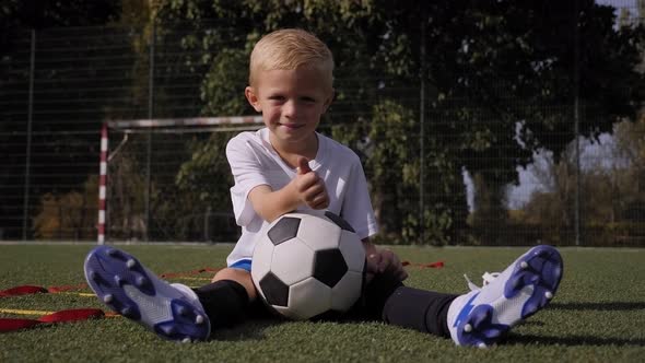 A Little Boy on the Football Field He is Sitting on the Grass with a Ball alt