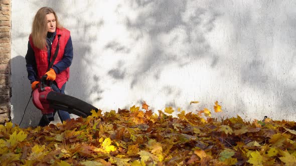 Female caucasian worker use electric powered leaf blower to blow autumn leaves