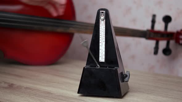 A black vintage metronome beats the rhythm while standing on the table, a cello is lying behind
