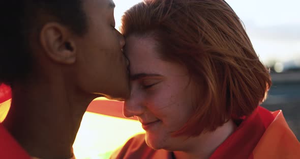 Multiracial lesbian girls having tender moment outdoor wearing LGBT rainbow flag alt