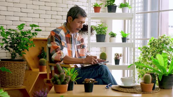 An old man sitting in a living room decorated with flower pots. alt