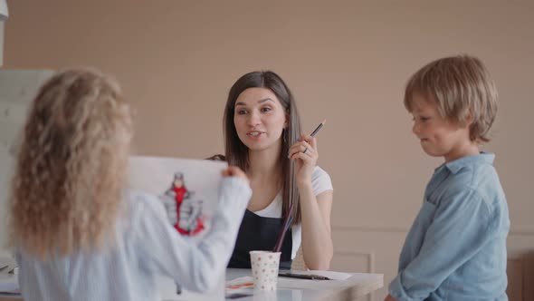 Group of Kids and Their Teacher Leaning Over Table with Watercolor Paints and Painting Selfmade Clay alt
