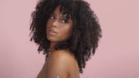 Mixed Race Black Woman with Curly Hair Covered By Crystal Makeup on Pink Background in Studio