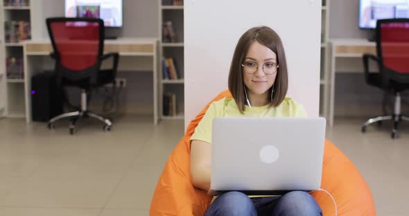 Young Woman is Smiling Sitting in a Beanbag Chair with a Laptop Working in the Library alt