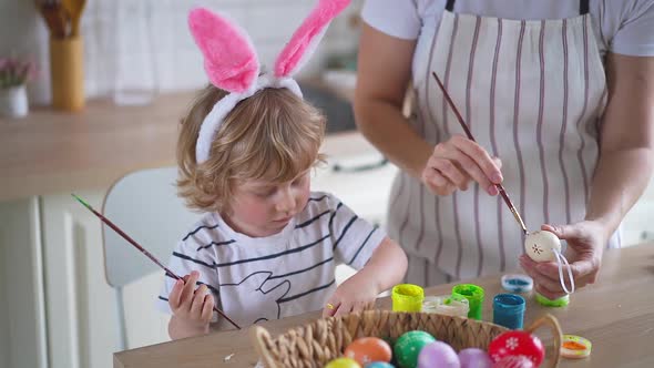 charming woman and her two-year-old son in bunny ears paint Easter eggs with paints