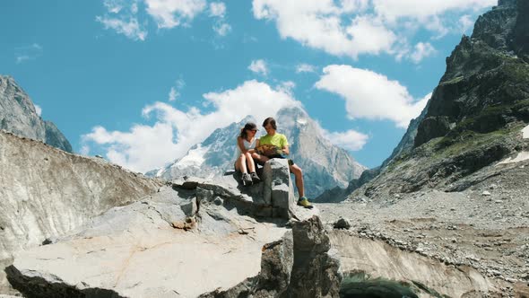 Couple of Tourists Sit on a Rock and Study a Map, Plan a Route in a Mountain Hike alt