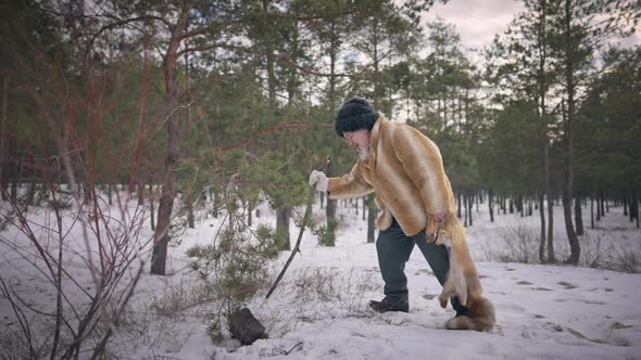 Indigenous Senior Hunter Standing in Winter Forest with Fox Skin Trying Snow with Stick alt