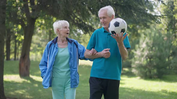 Front View Dolly Shot Happy Fit Senior Man and Woman Walking Arm in Arm with Soccer Ball Talking in alt