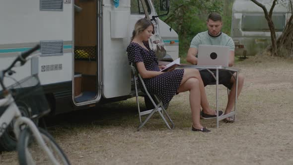 Man working at laptop and woman reading the book in summer camping. alt
