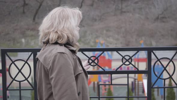 Side View of Thoughtful Senior Caucasian Woman Standing Next To Fence and Looking Away. Blond Mature alt