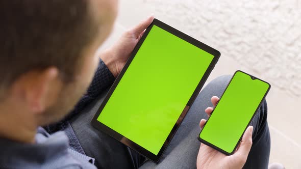 A Man Looks at a Smartphone and a Tablet (Both Vertical) with Green Screen  Closeup From Behind