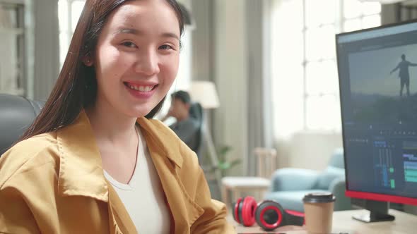 Asian Female Video Editor Works With Footage And Sound On Computer. He Turns And Warmly Smiles alt