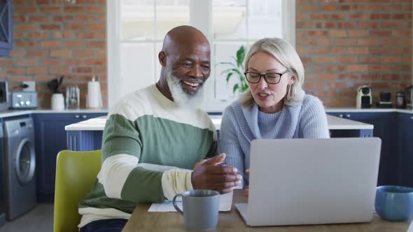 Happy senior diverse couple in kitchen sitting at table, using laptop alt
