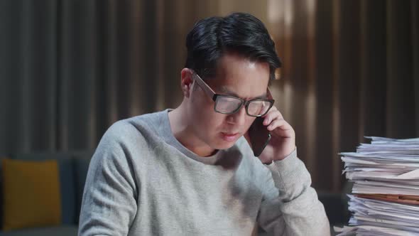 Close Up Of Asian Man Talking On Phone And Taking Note While Working Hard With Documents At Home alt