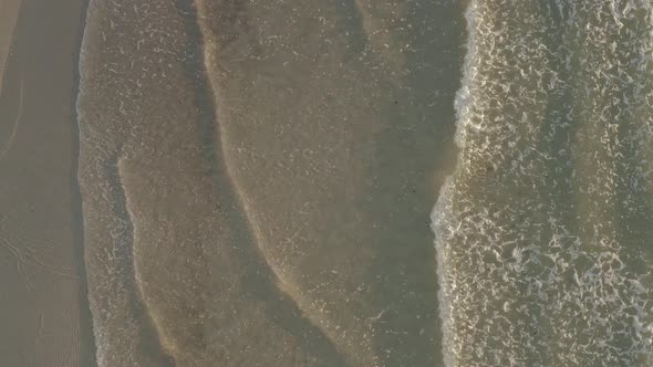 Aerial ascending slowly on rolling waves breaking on sandy sunlit beach alt
