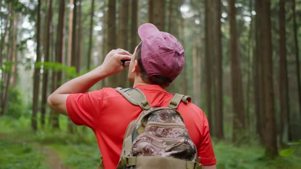 Young Male Tourist with a Backpack Drinks While Walking in the Forest, Camera Tracking, Back View alt