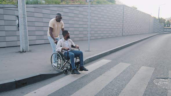 African American Adult Man Crossing Road at Crosswalk Pushing Young Bearded Guy on Wheelchair alt