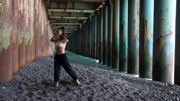 a Barefoot Woman Dances Under the Pillars of the Bridge Against the Background of the Incoming Waves alt