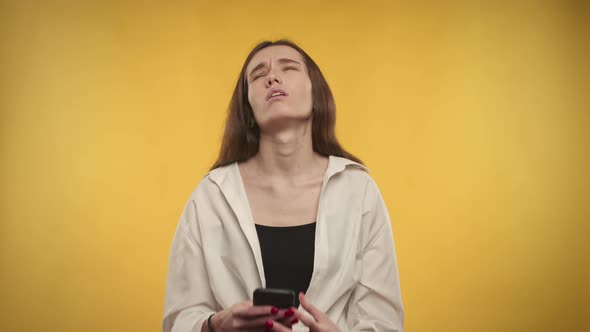 Young Adult Caucasian Woman is Upset By the News on Her Smartphone on a Bright Yellow Background alt